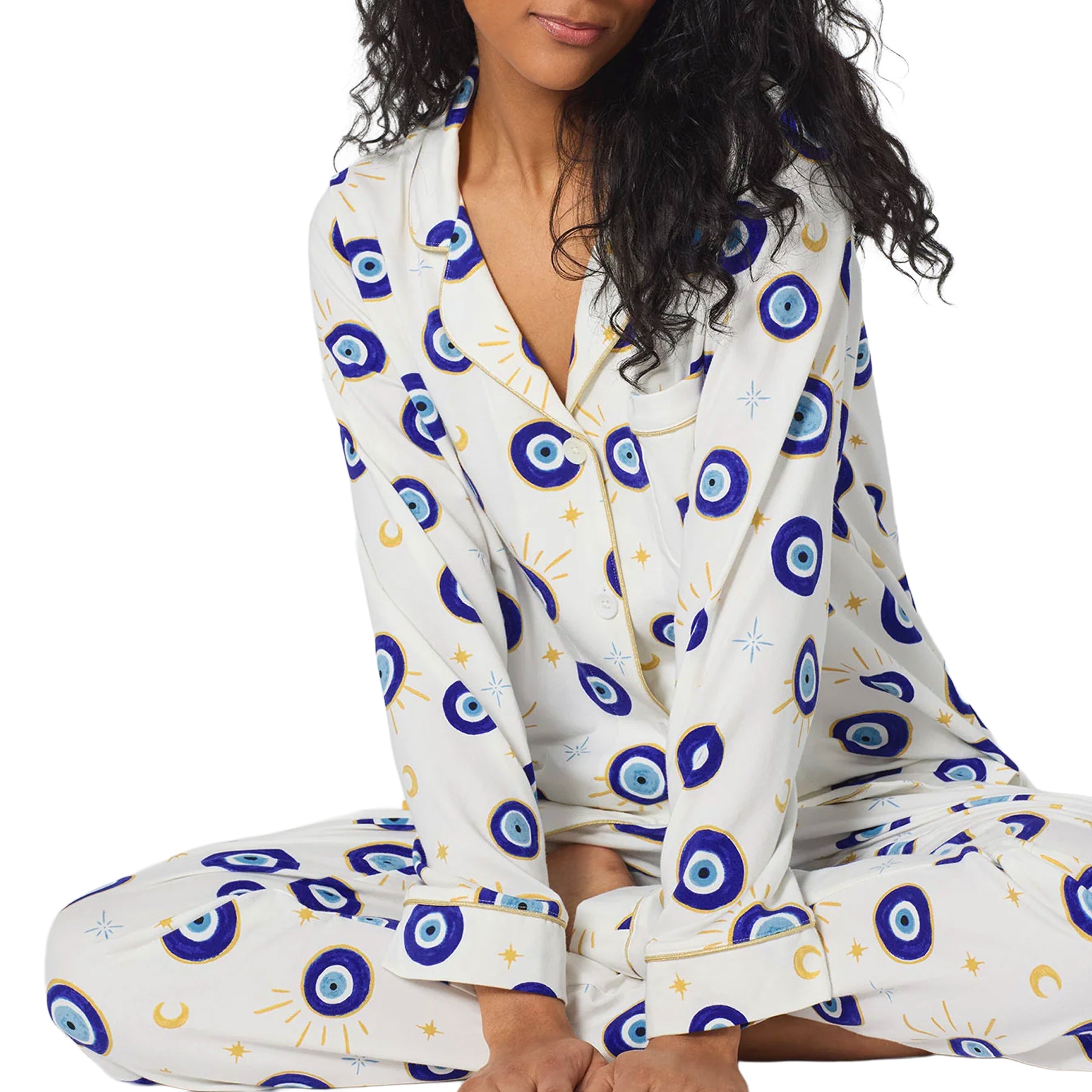 Woman wearing a white BedHead classic pajama set with blue evil eye, moon and star print, shown seated.