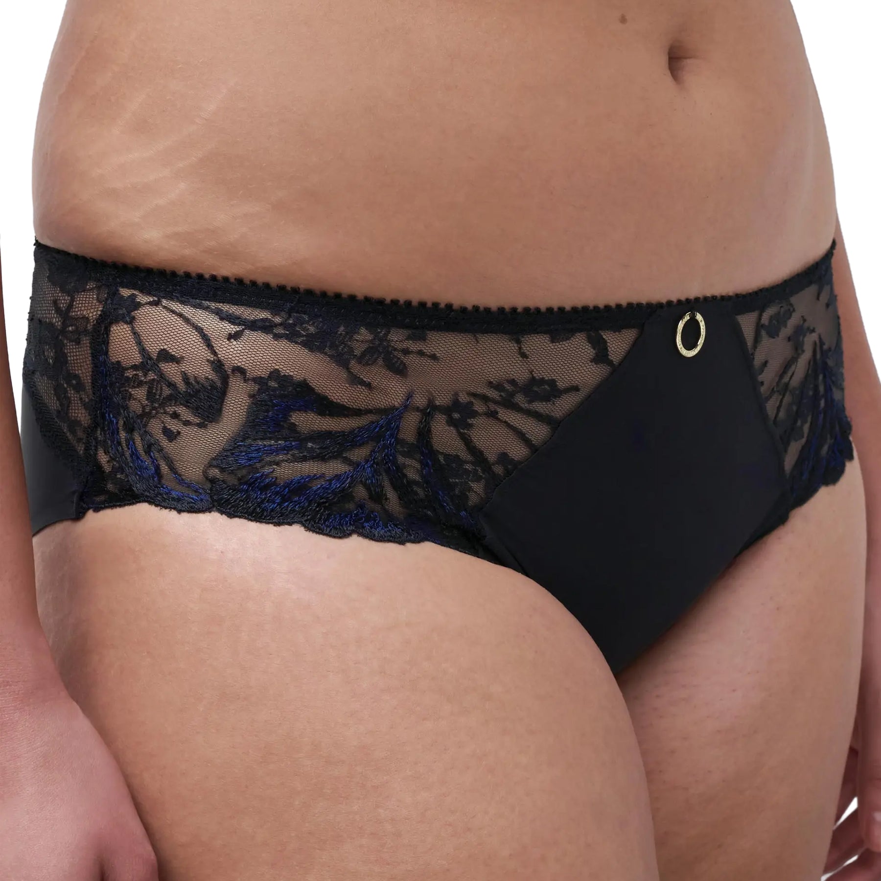 Model wearing a black full brief with sheer embroidered floral side panels and gold-tone ring accent.