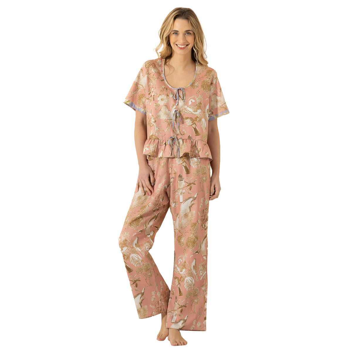 Woman modelling a dusty pink bird and floral print pajama set featuring a ruffled hem top, lavender tie details and relaxed full-length pants.