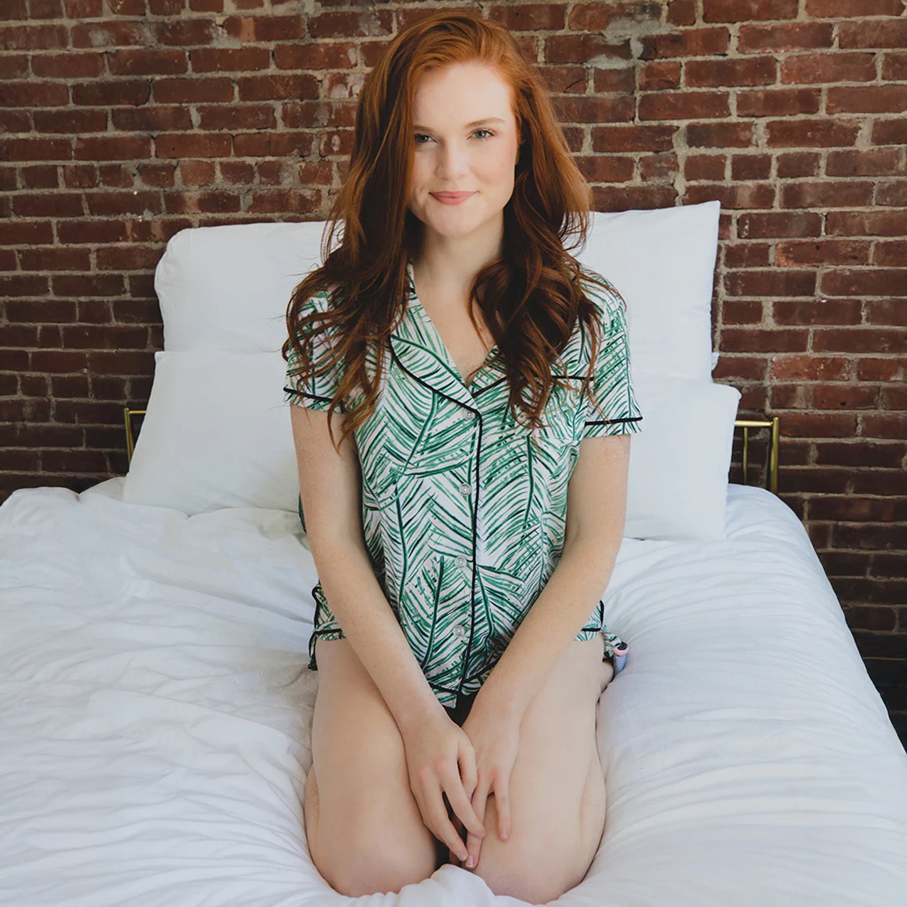 Model sitting on bed wearing green palm print pyjama set with short sleeve button-up top and matching shorts.