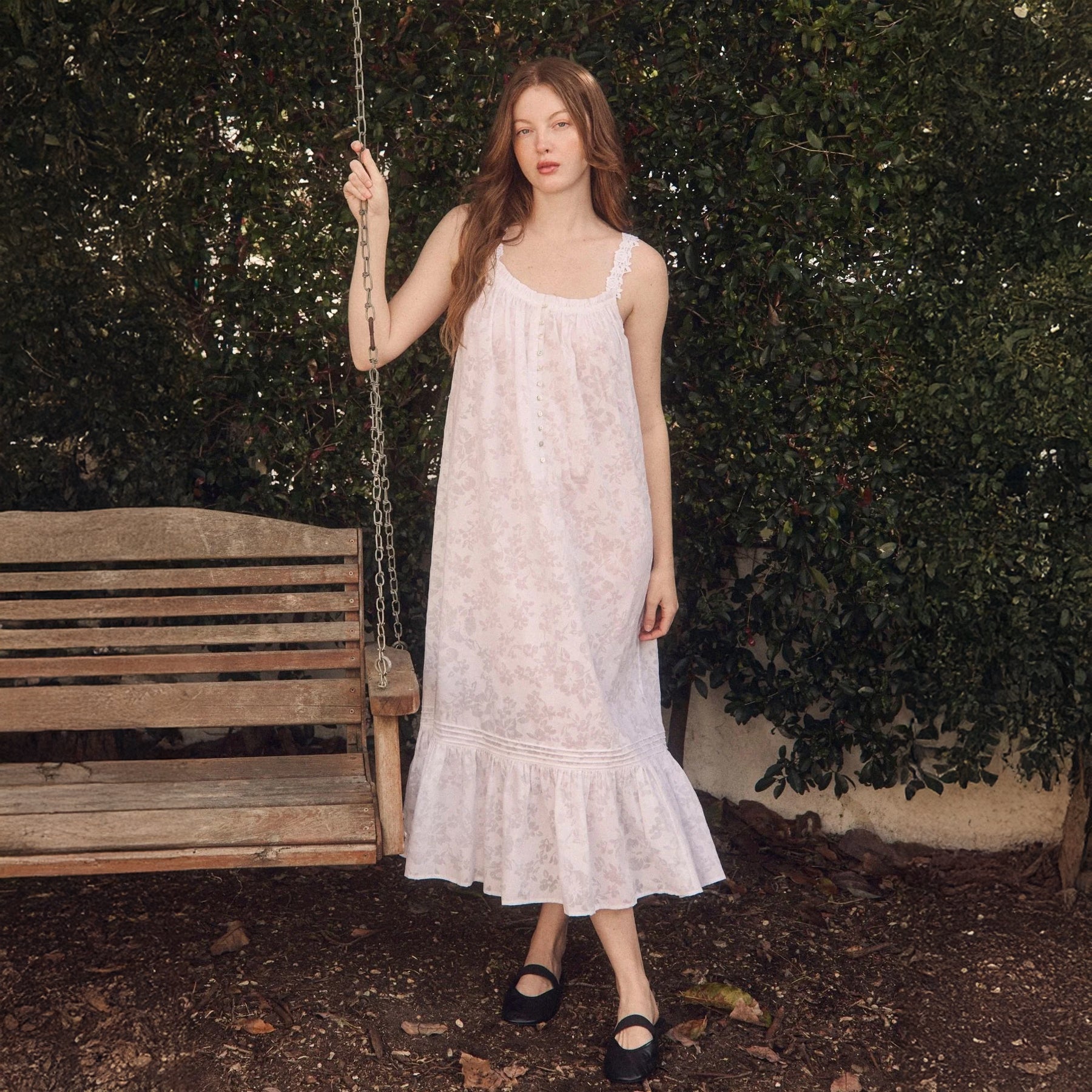 Woman standing outdoors by a wooden swing wearing a soft cotton nightgown in pale blush floral print with lace straps and ruffle hem