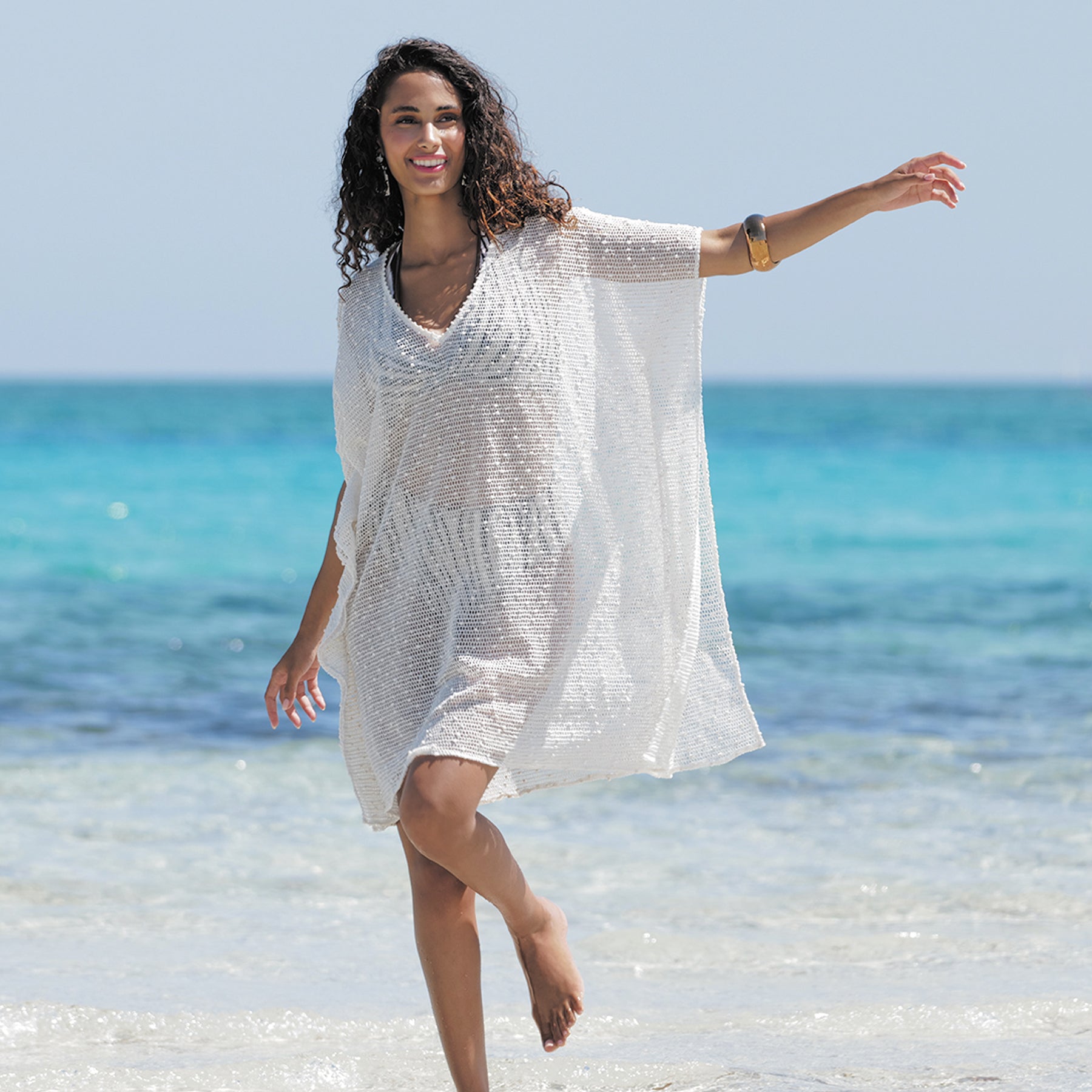 Woman in a white cover-up standing on a beach with clear blue water and sky.