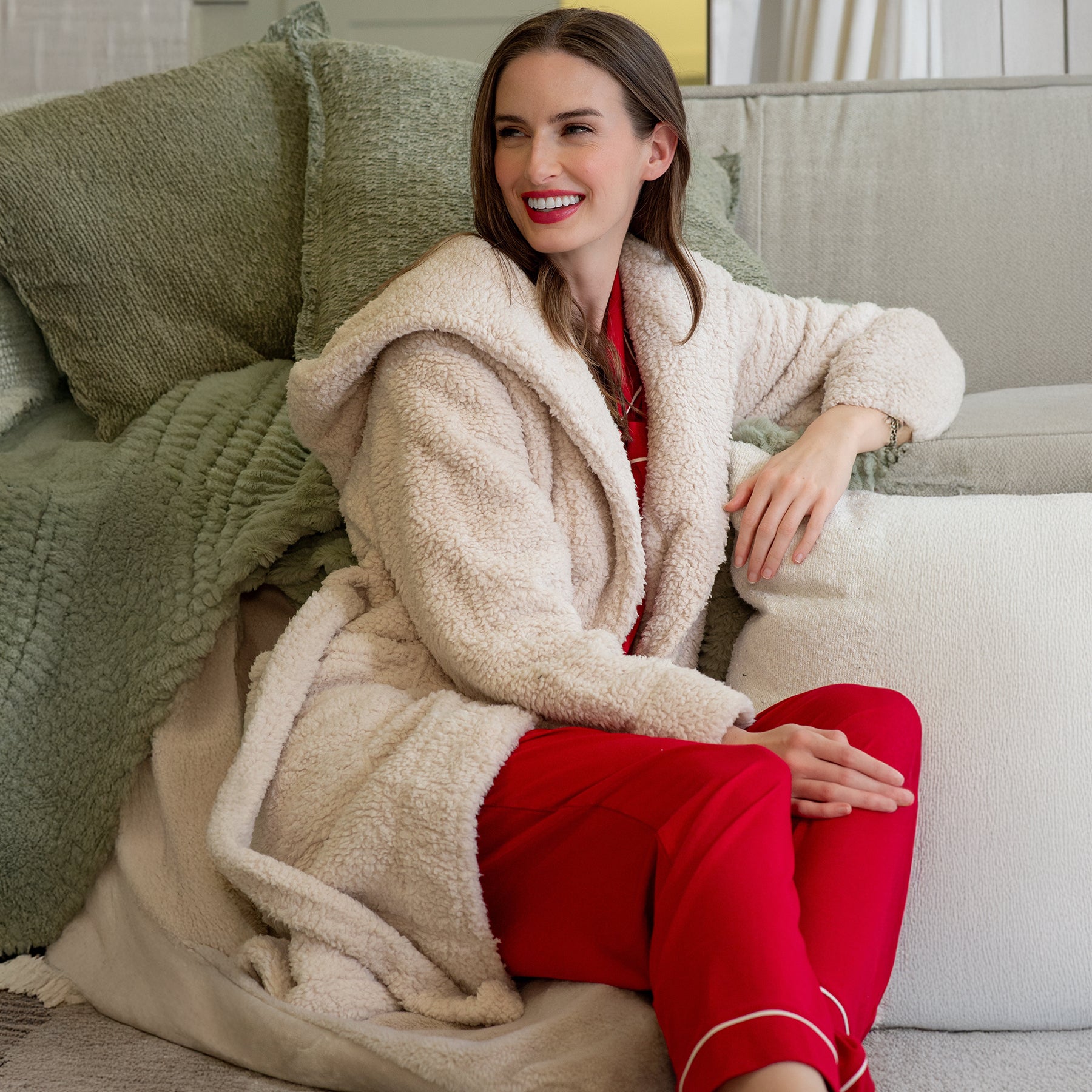 Woman sitting on sofa wearing LatteLove Sherpa Robe with Hood in Toasted Marshmallow, layered over red pyjamas.