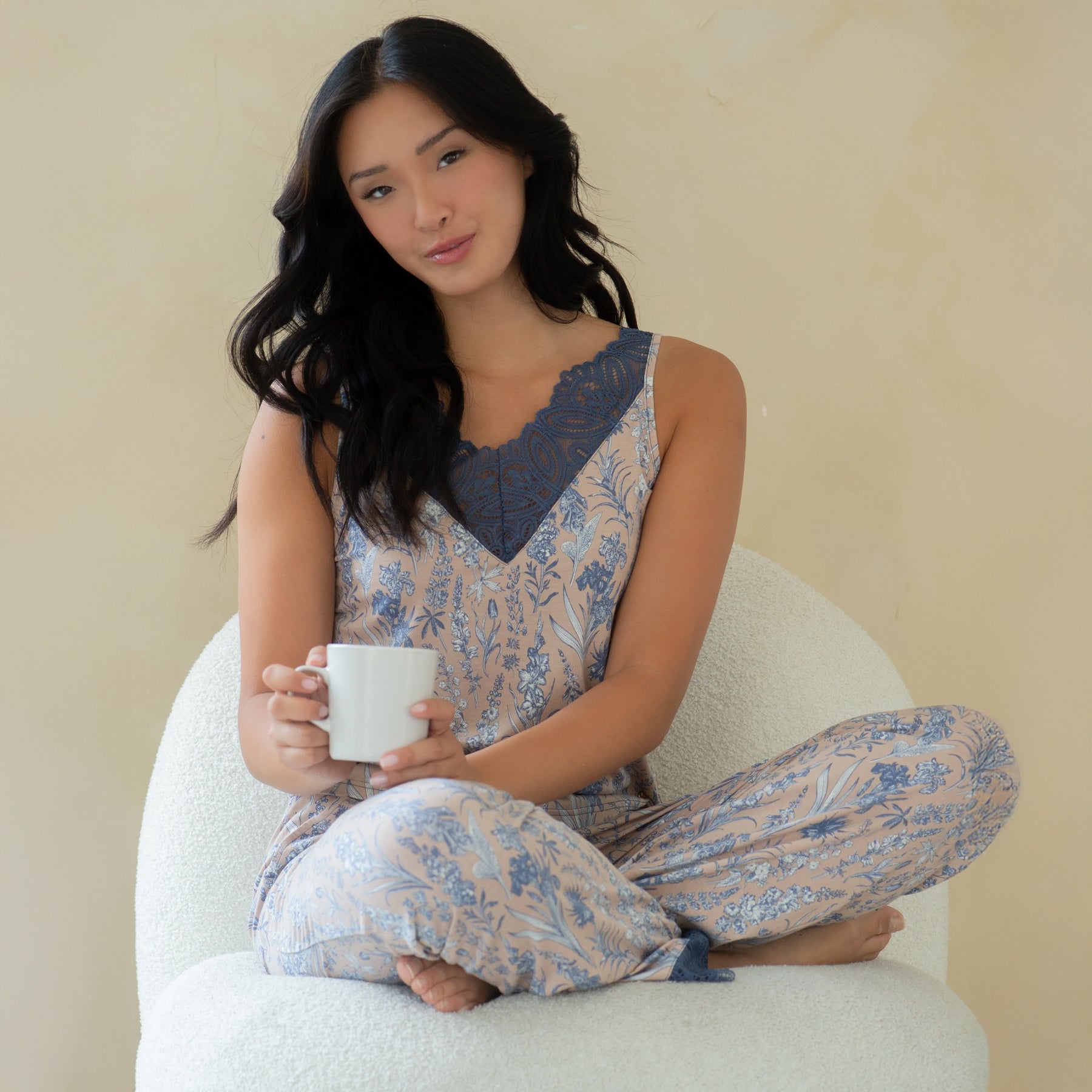 Woman sitting in a chair wearing a beige and blue floral print pajama set with lace neckline, holding a cup in a relaxed setting