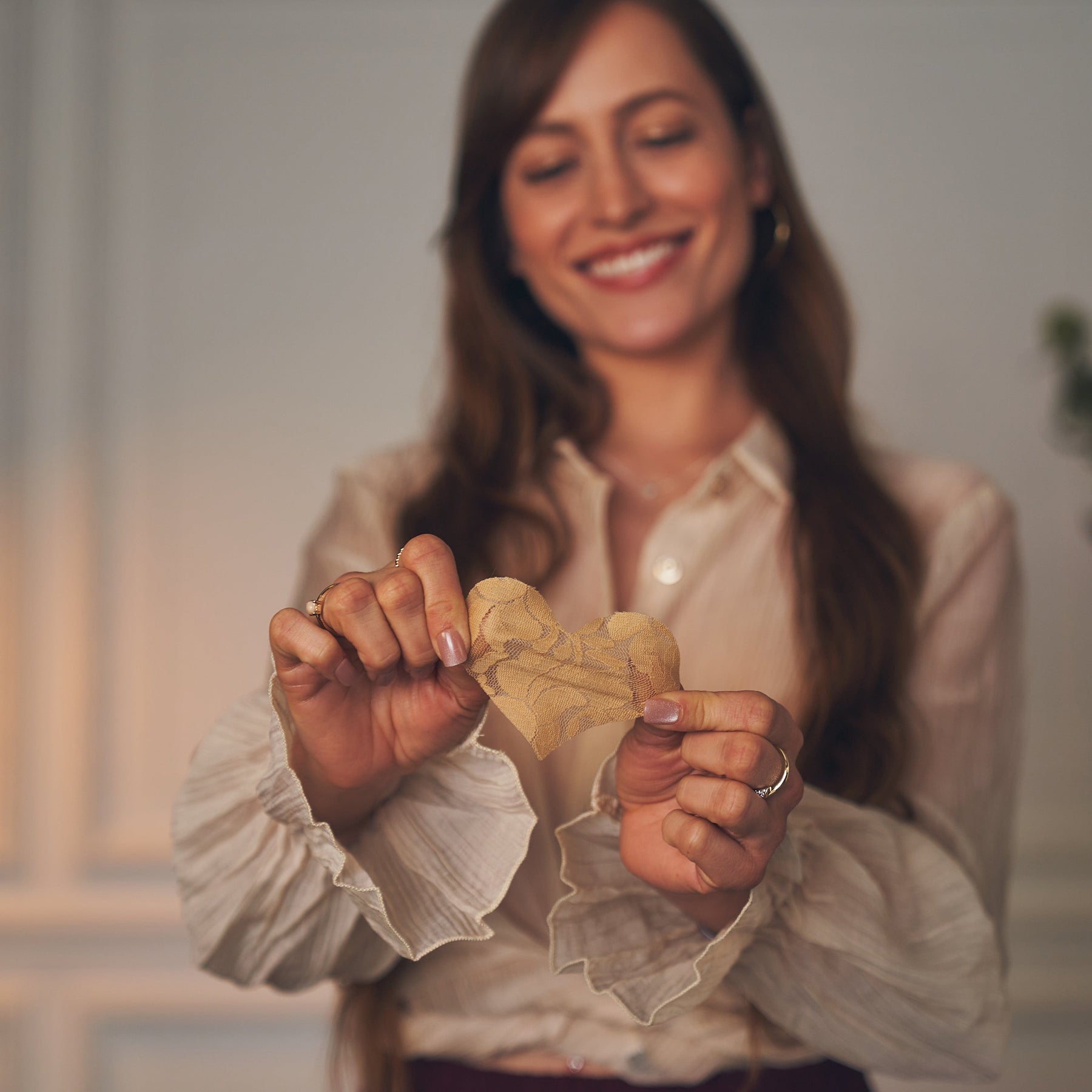 Woman holding up lace heart-shaped Nippies nipple cover with a smile, wearing a light blouse