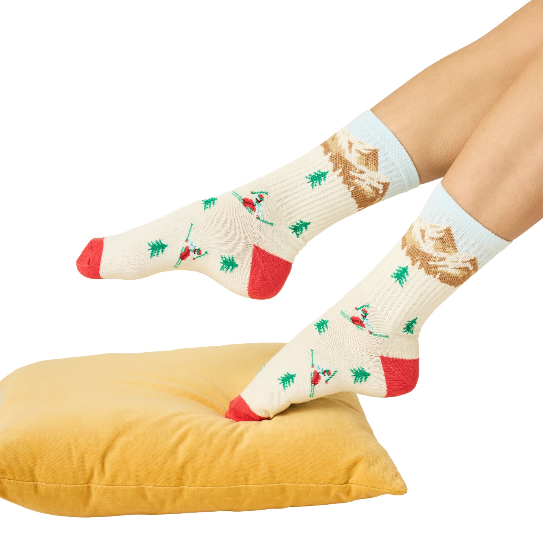 Ivory skiing-themed socks with mountain, pine tree and skier motifs, styled on feet over a mustard yellow cushion.