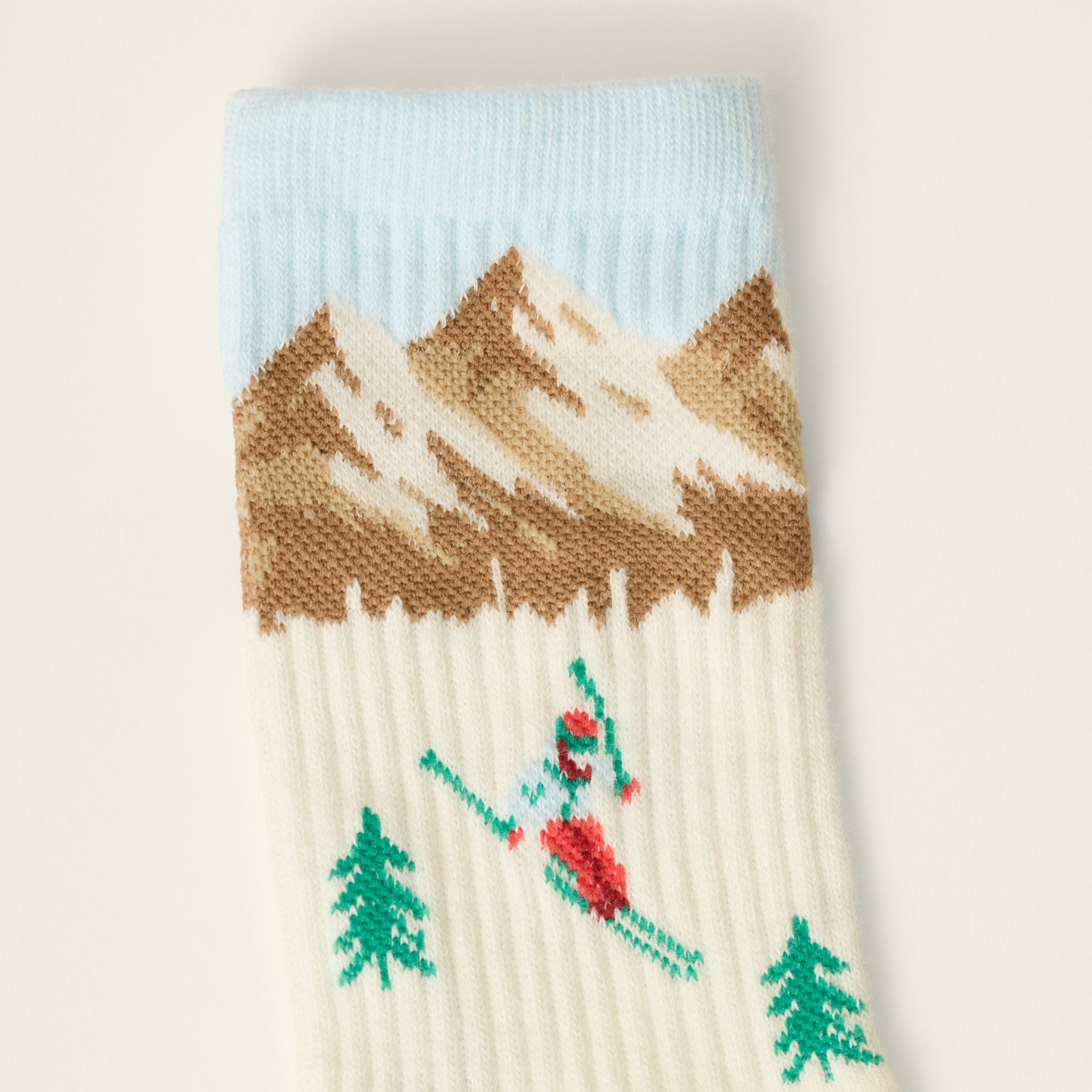 Close-up of ivory knit sock featuring a skier, green pine trees, snow-capped brown mountains and a light blue cuff.