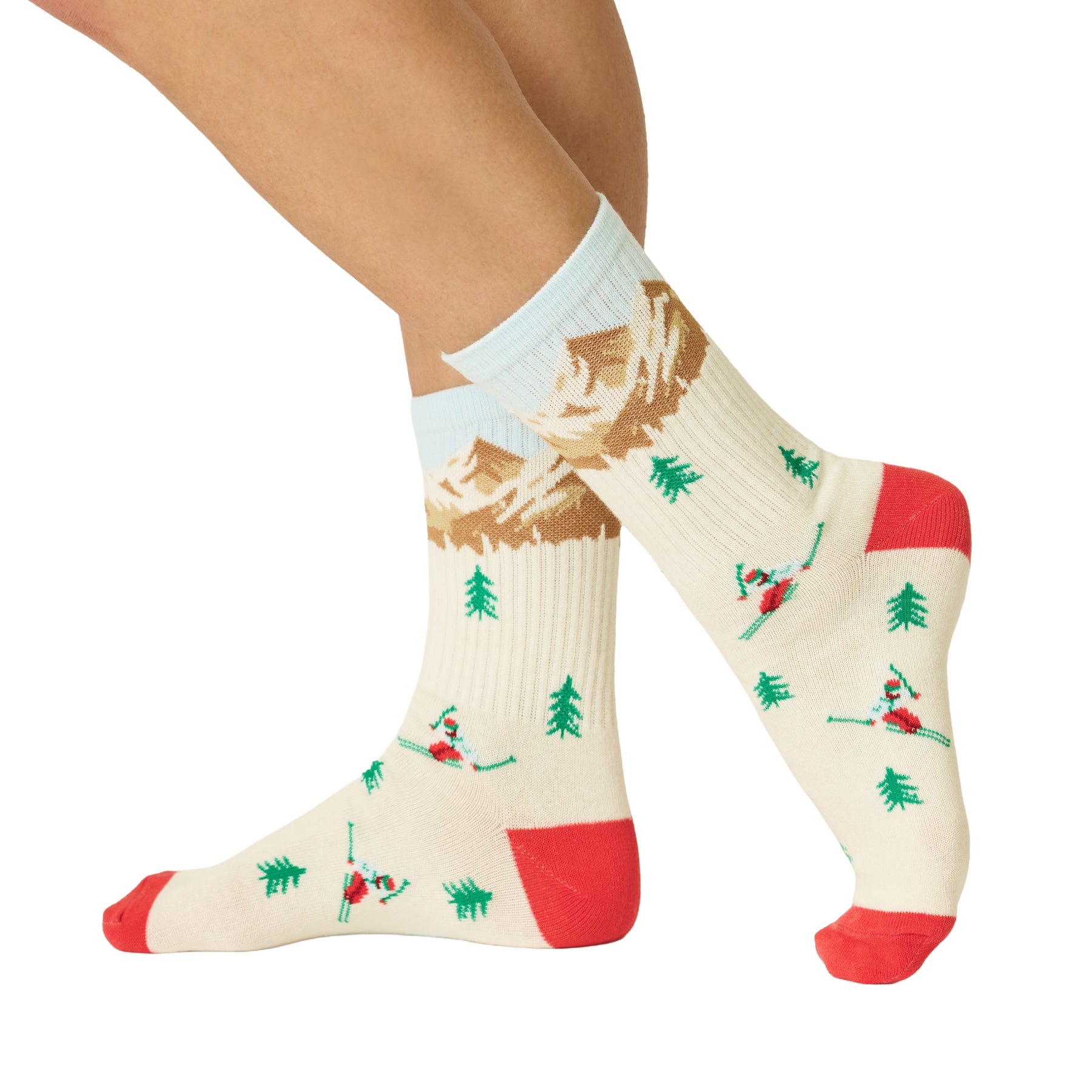 Side view of ivory skiing socks featuring snow-capped mountains, skiers, green pine trees, red toes and light blue cuffs.