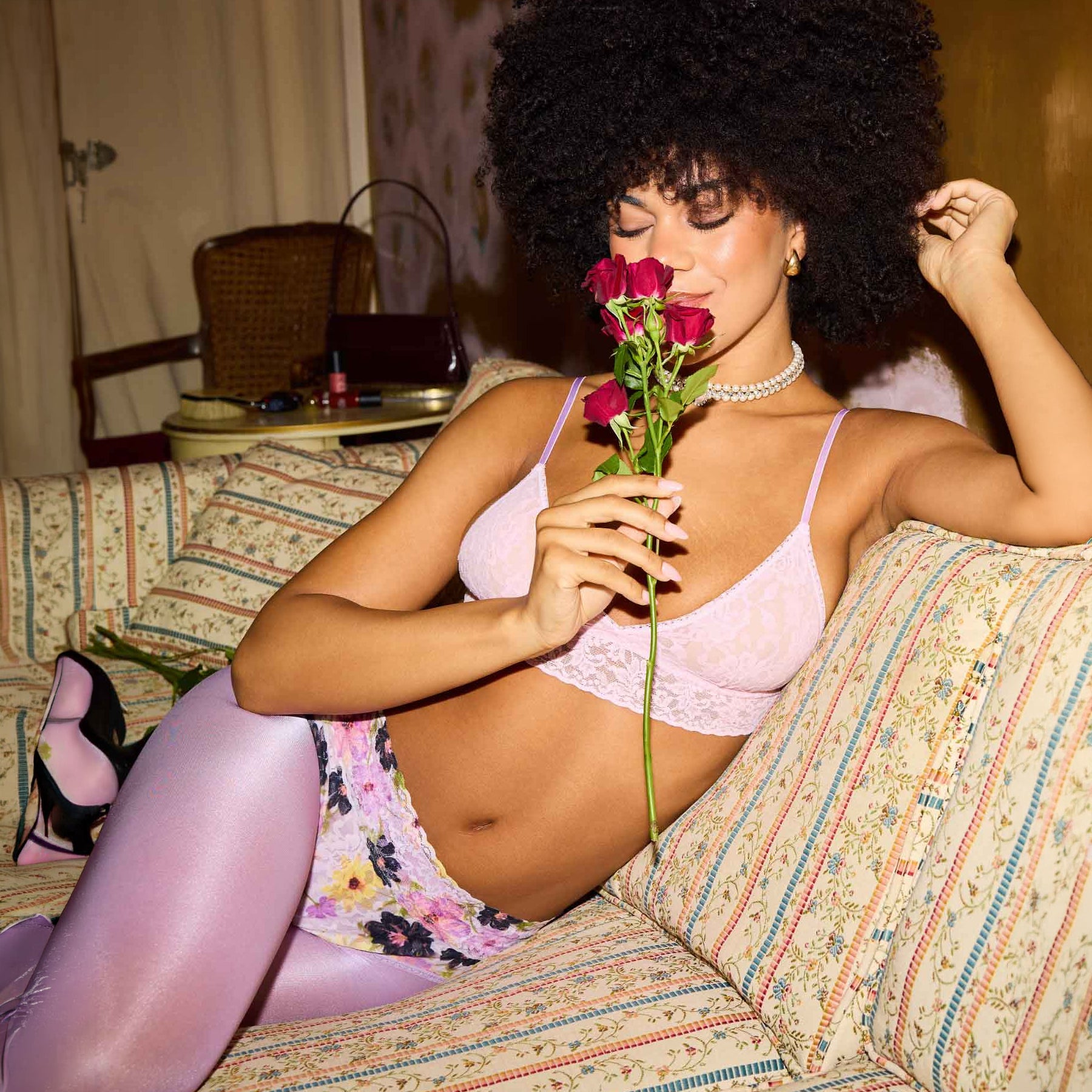 Model relaxing on a patterned sofa wearing a floral lace high-waist brief and light pink lace bra, holding red roses.