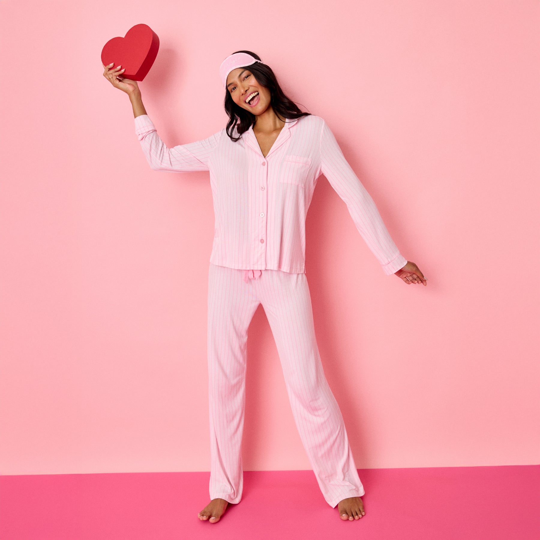 Woman wearing pink striped pyjama set holding red heart, soft loungewear styled for Valentine’s theme