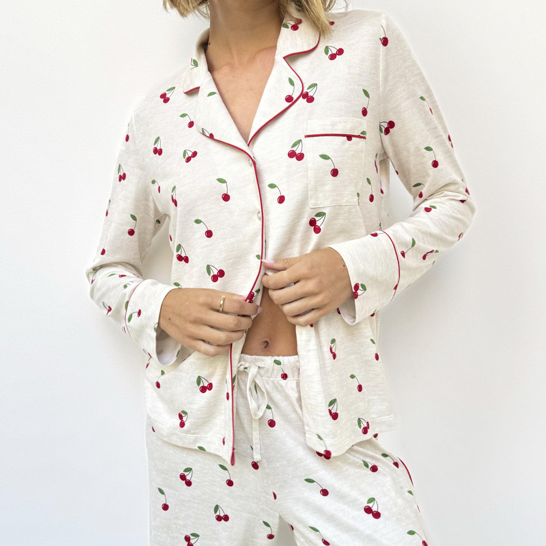 Woman wearing beige pyjama set with cherry print and red piping, buttoning the top.