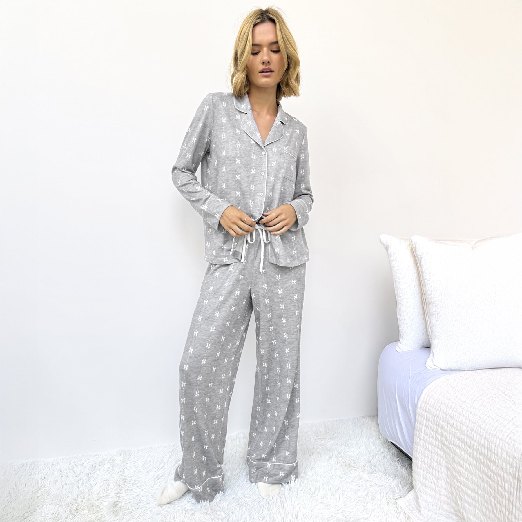 Woman wearing grey pyjama set with white bow print and white piping, standing beside bed.