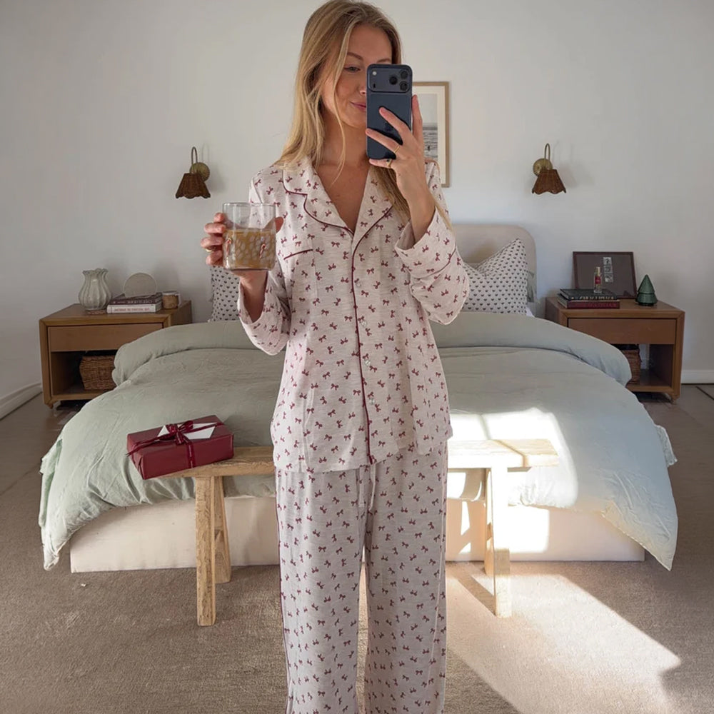 Model wearing Splendid Pillowsoft white bow pajama set with button-up top and matching drawstring pants, holding a drink in a bedroom.
