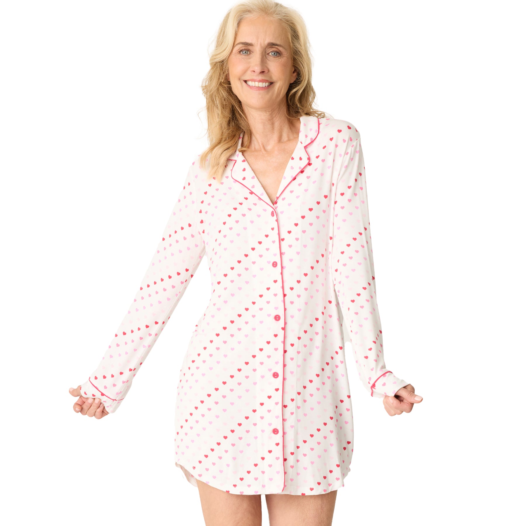 A woman wears a long-sleeve white pajama shirt with a heart print and pink piping, smiling comfortably.