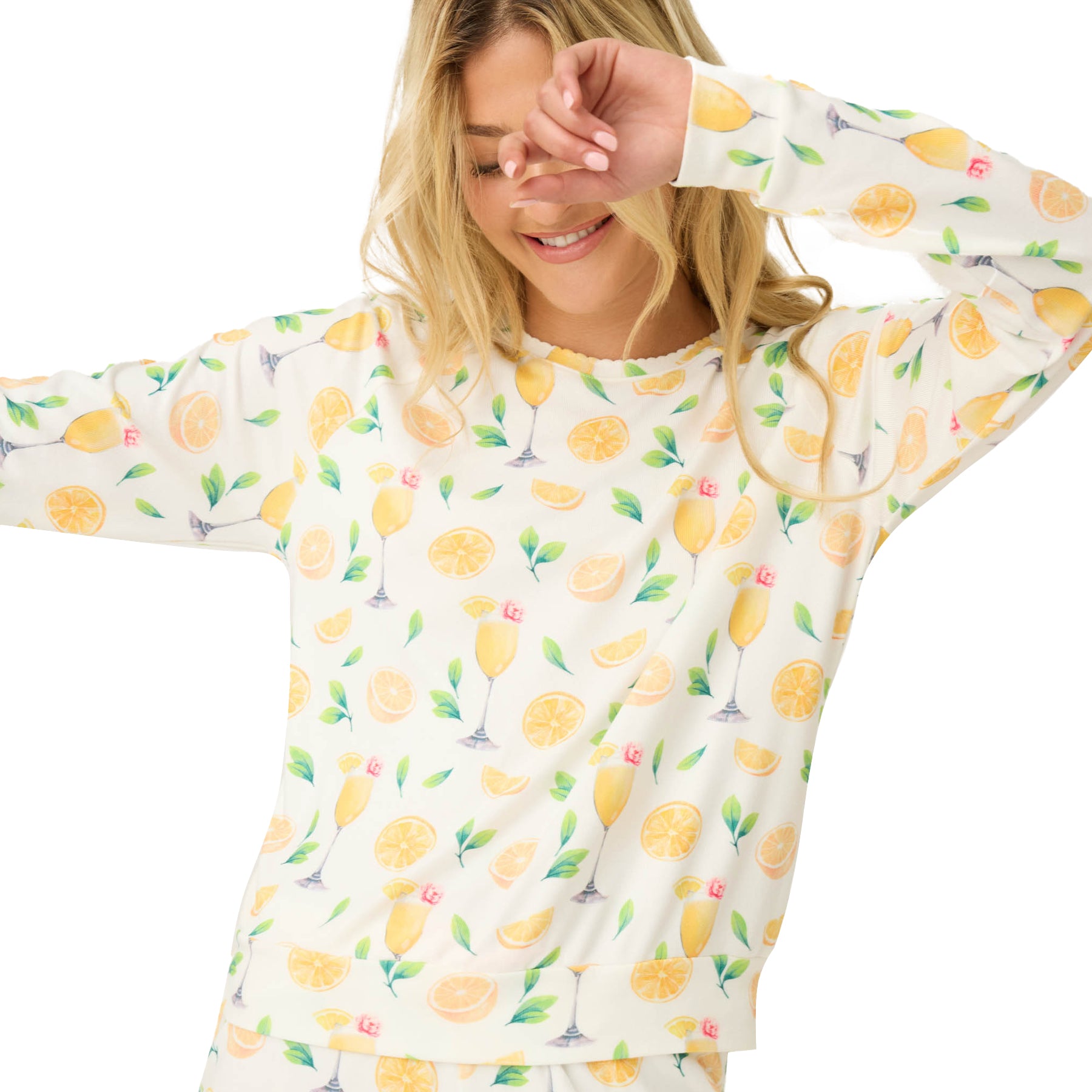 Smiling woman in a long-sleeve ivory pyjama top with a lemon and mimosa cocktail print, playfully covering part of her face with her sleeve.