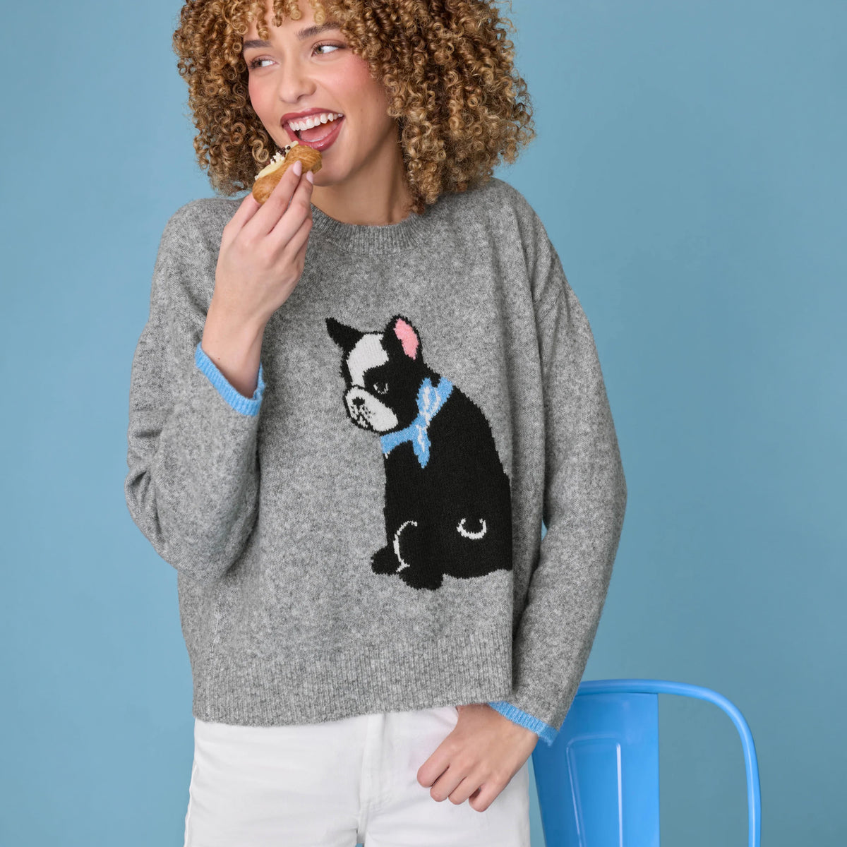 A woman with curly hair wearing a gray knit sweater featuring a black and white French Bulldog graphic with a light blue bow tie. She is holding a pastry near her mouth, wearing white pants, and standing in front of a solid blue background.