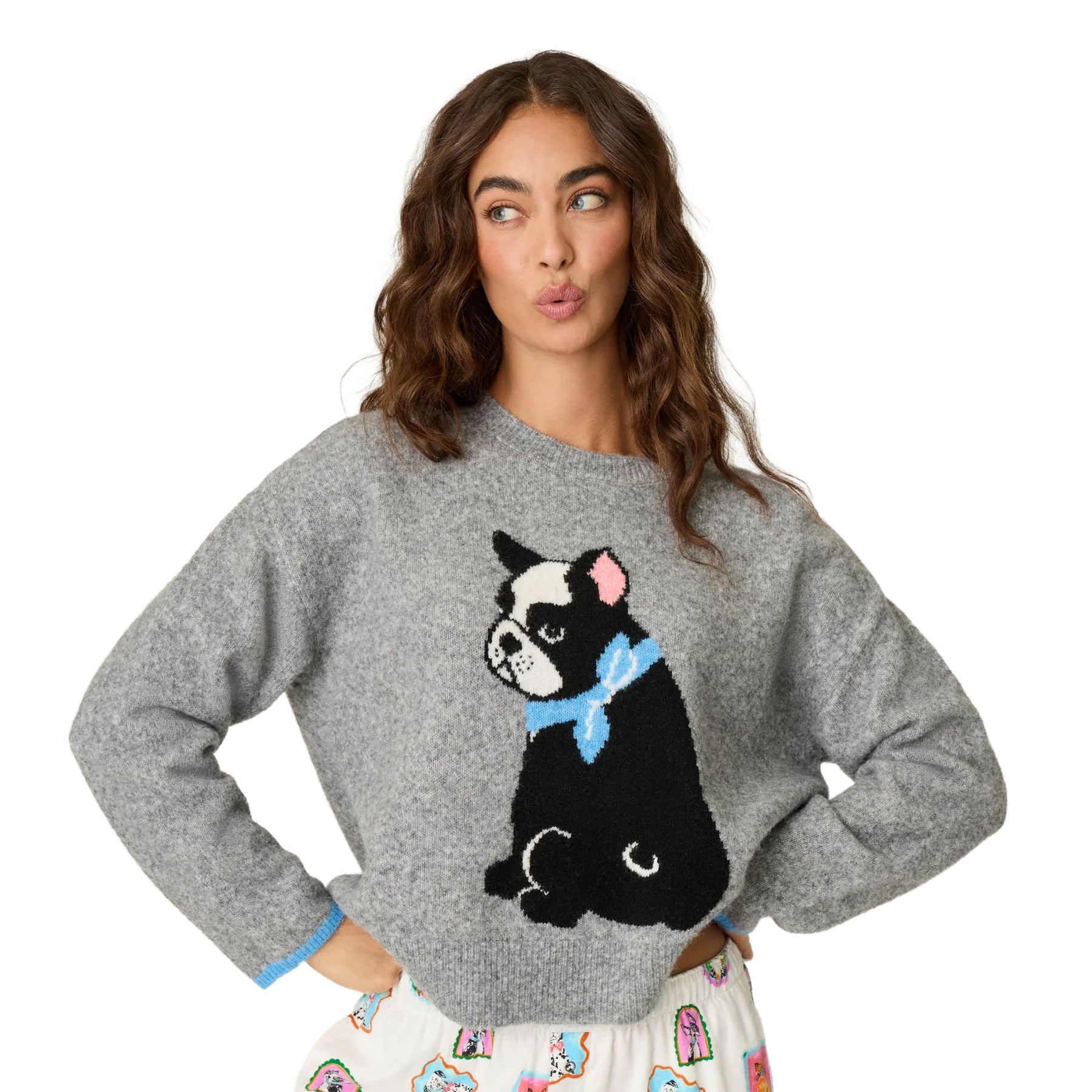 A woman modeling a gray knit sweater featuring a large black-and-white dog graphic with a light blue bow tie on the front, paired with a white patterned skirt against a plain white background.