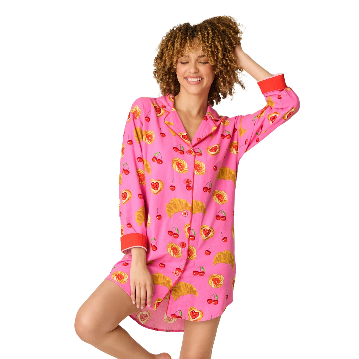 Model wearing a bright pink long-sleeve sleep shirt featuring a playful cherry, croissant and pastry print with red contrast cuffs.