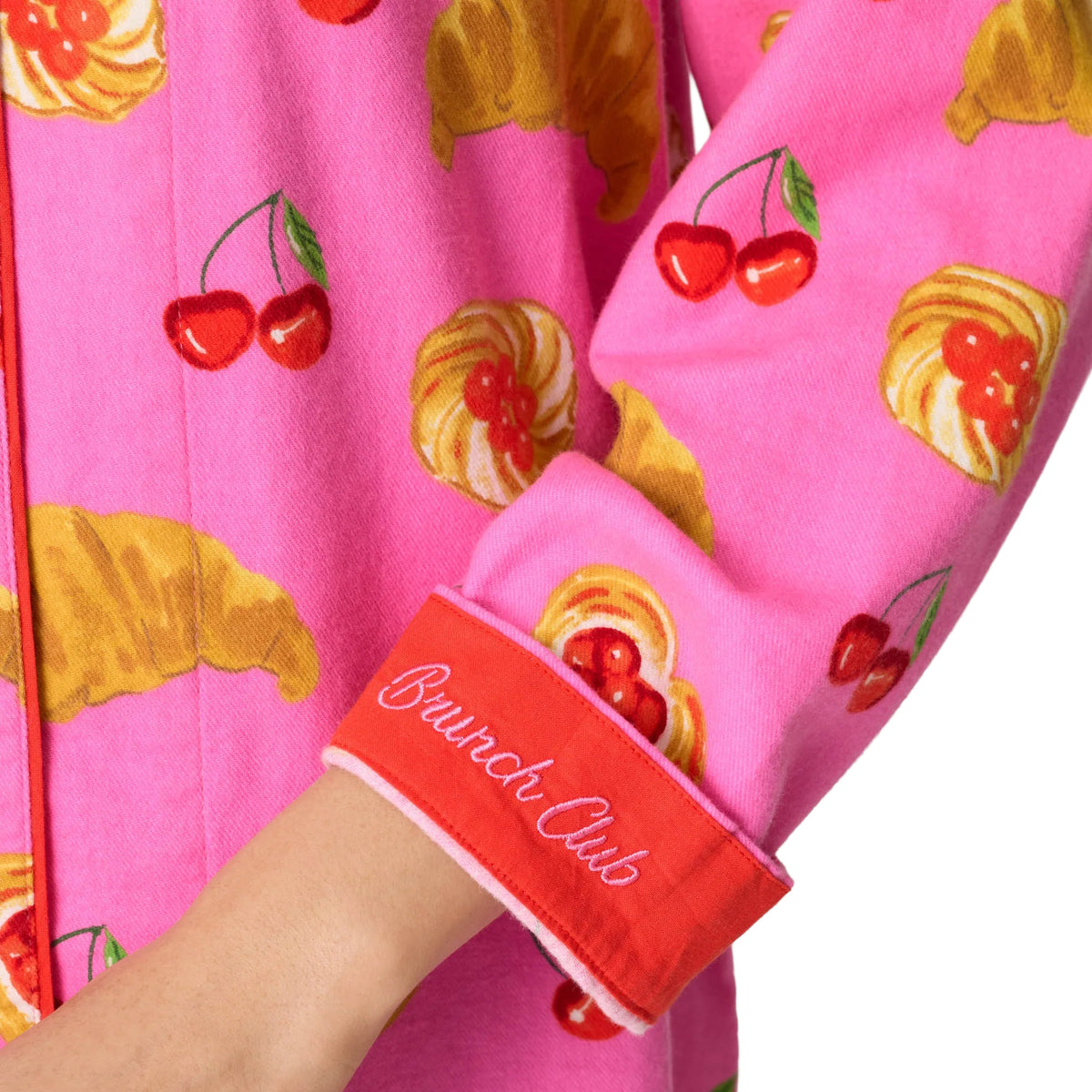 Close-up of a person wearing a bright pink long-sleeve top with a print of red cherries and yellow croissants, featuring a red cuff with white script that reads 'Brunch Club'.