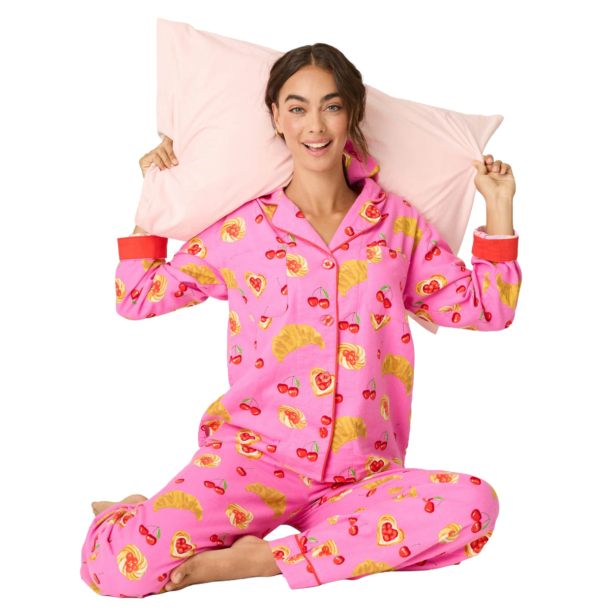 Woman sitting cross-legged in bright pink pajamas featuring a playful cherry, croissant and pastry print with red trim.