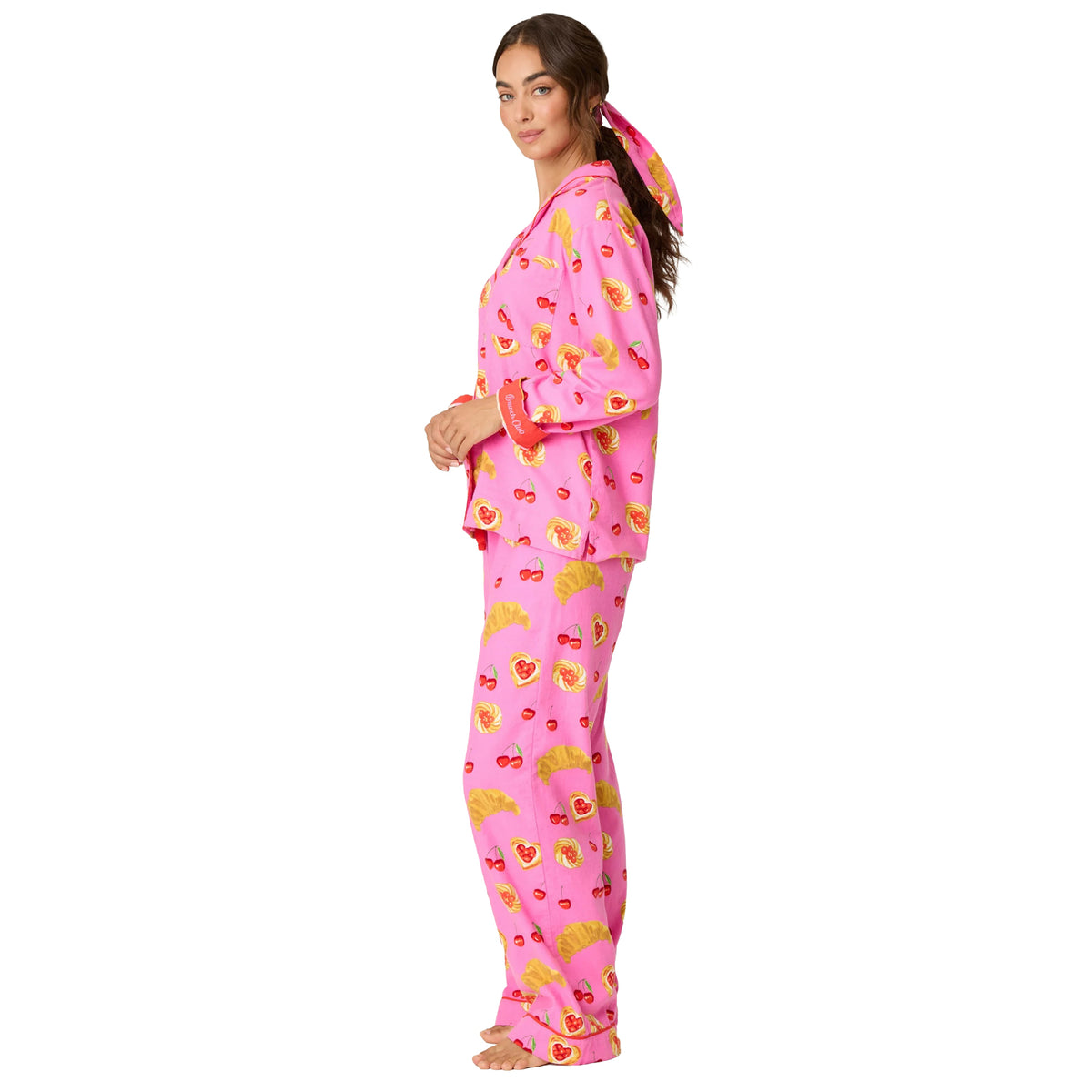 Side view of woman wearing bright pink cherry and croissant print pajamas with red cuff detailing and matching hair accessory.
