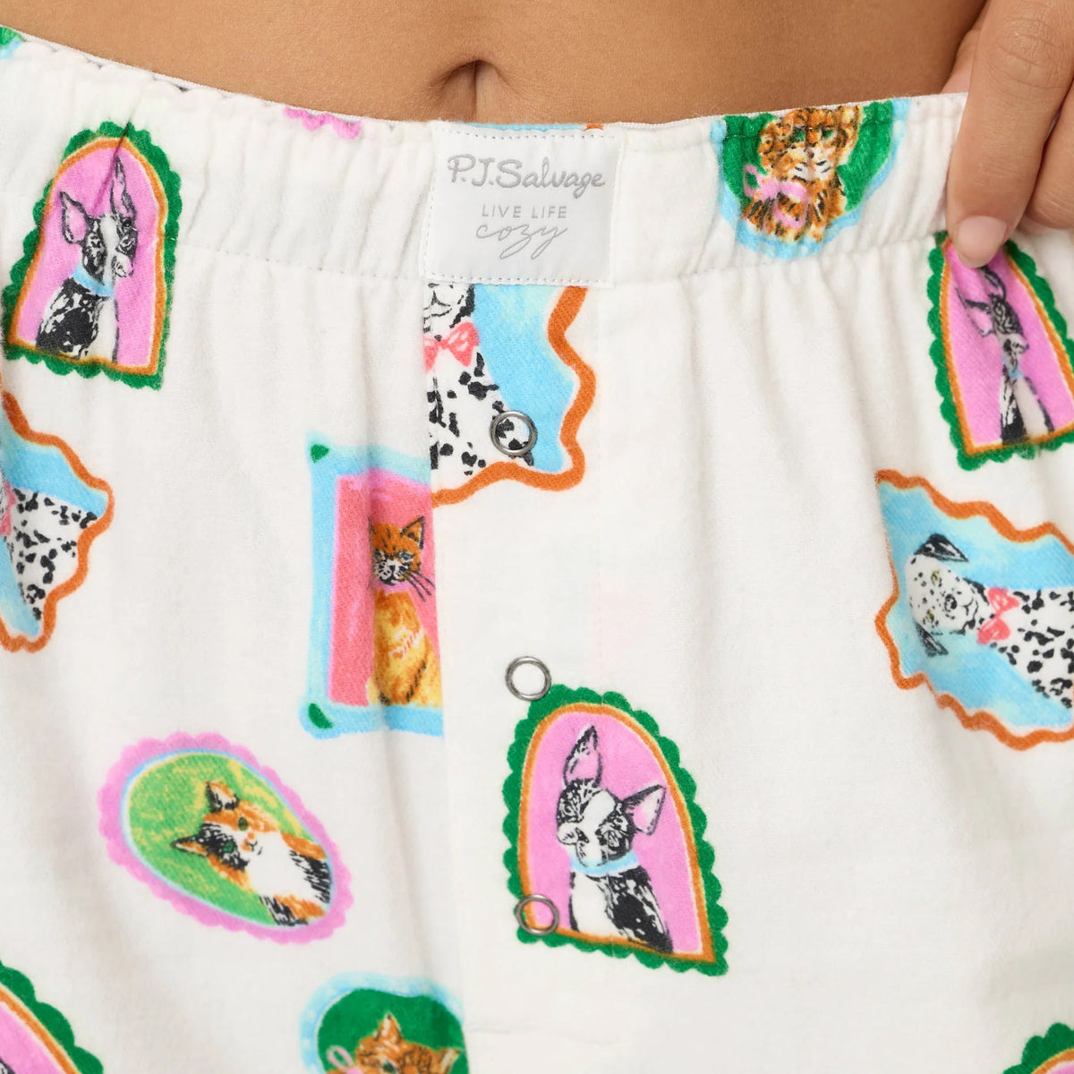 Close-up of cream pajama pants showing the elastic waistband, button fly and colorful framed dog and cat print.