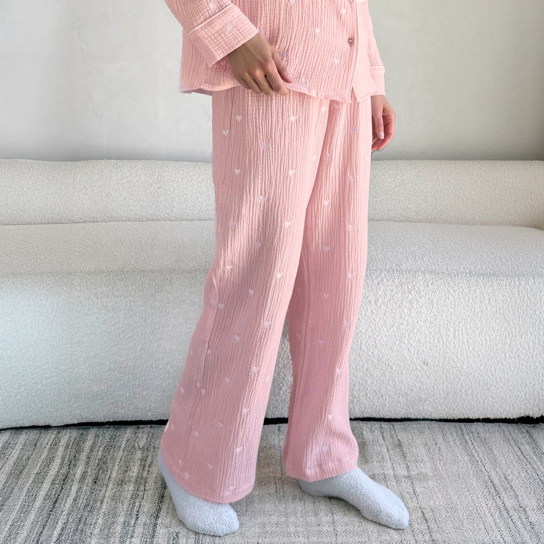 classic button up pyjama set in crinkle cotton in baby pink with mini white embroidered hearts. Matching white  contrast piping on trims. Close up view of pants on model.