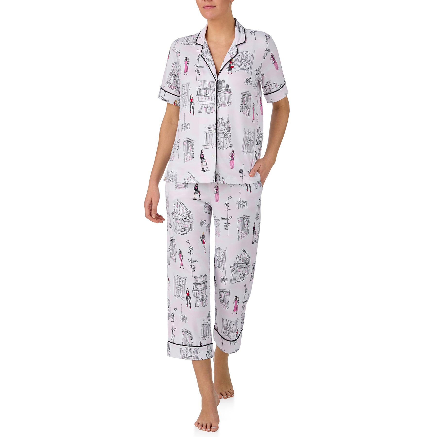 Woman wearing a white city-print pyjama set with a short sleeve button-up top and cropped lounge pants featuring illustrated fashion sketches and Paris-inspired scenery.